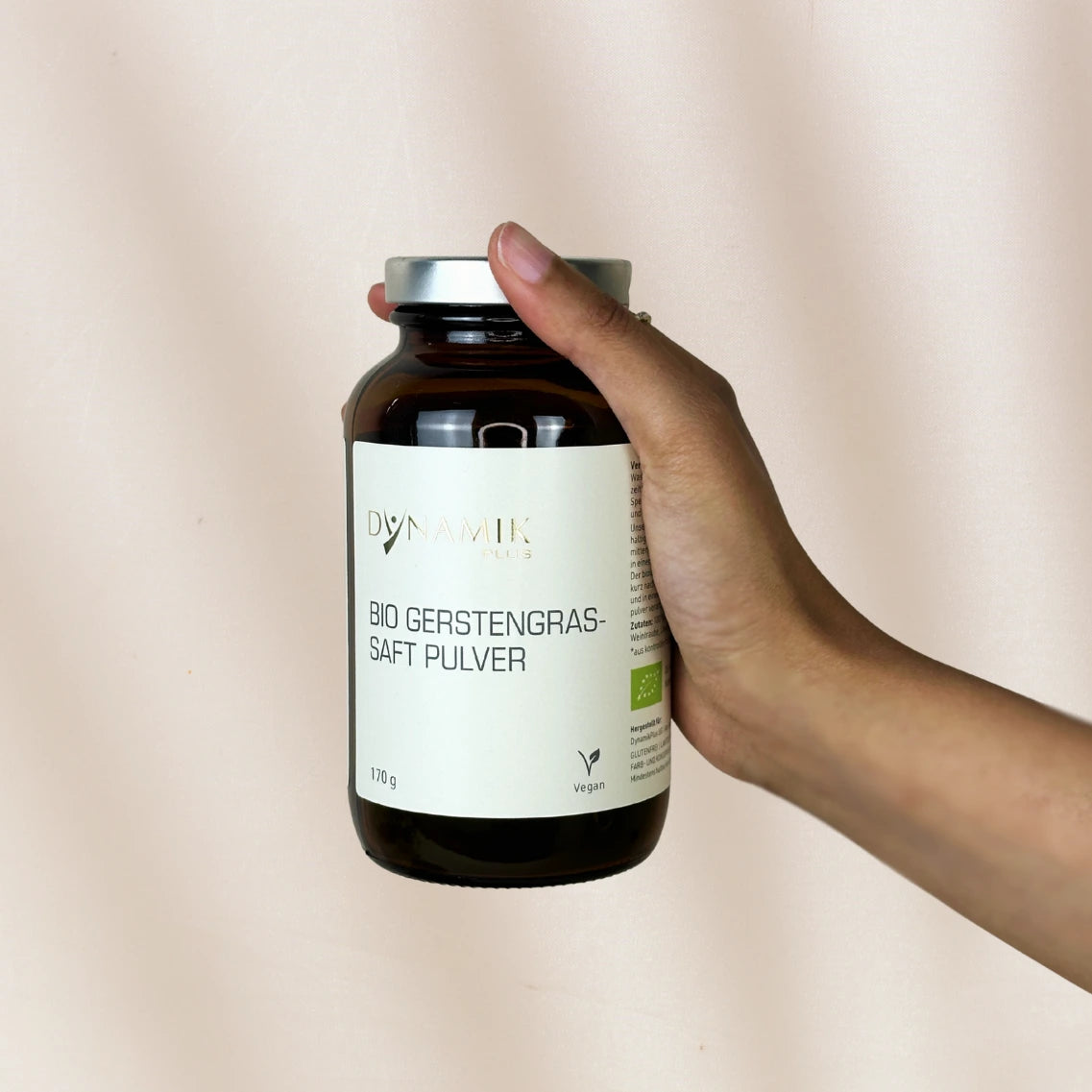 Braune Glasbehälter mit DYNAMIKPLUS "Bio Gerstengrassaft Pulver" Etikett von Hand gehalten