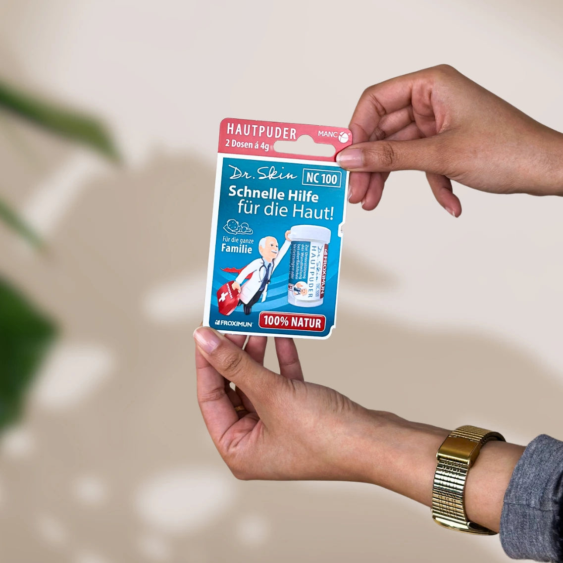 Blaue Verpackung mit "Dr. Skin Hautpuder" Druck und Arztabbildung, gehalten von zwei Händen