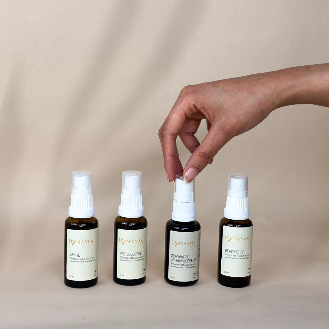 Vier braune Sprühfläschchen mit weißem Sprühkopf und DynamikPlus Etikett, eins wird gehalten von einer Hand