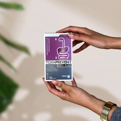 Rechteckige violet-weiße Verpackung mit "TOXAPREVENT Medi Plus mit Zeolith" Print und Verdauungstraktabbildung in Hand gehalten