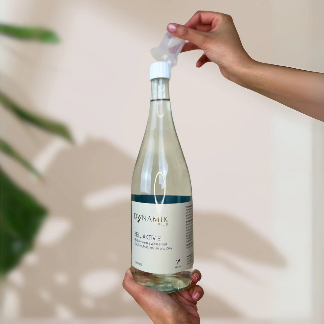 Glasflasche mit weißem Deckel, kleinem Messbecher und DynamikPlus "Zell Aktiv 2" Etikett gehalten von zwei Händen